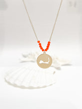 Load image into Gallery viewer, the beach and back cape cod necklace in gold with coral beads at center of chain and coin shape pendant with cutout silhouette of cape cod pictured in front of white seashells on white background